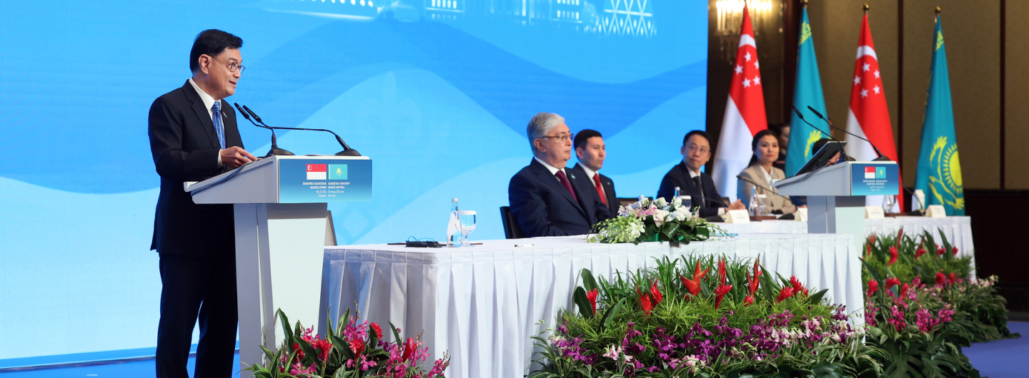Conference with speaker at podium, flags of Singapore and Kazakhstan, and seated dignitaries.